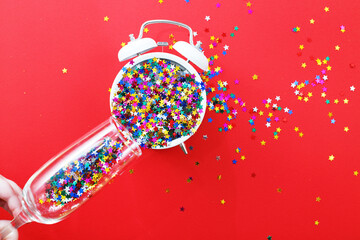 hands hold champagne glass with sequins pour on the alarm clock on a red background. christmas, celebration concept.
