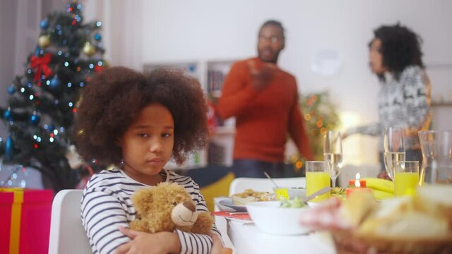 Sad girl with toy looking at camera, stressed parents fighting on background