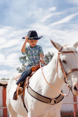 Kid in cowboy boots and hat riding a white horse on a ranch