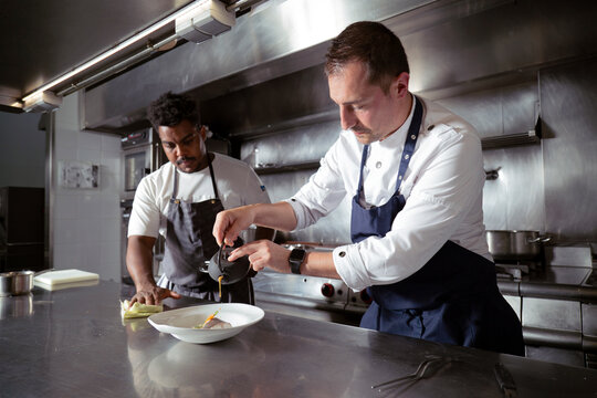Chef pouring bouillon to fish while working with black assistant