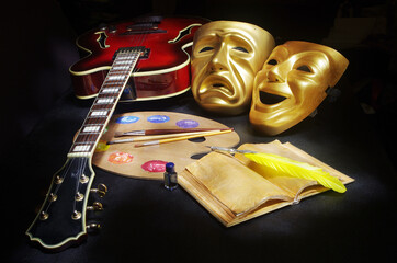 Attributes of the arts. Music, painting, theater, literature. Electric guitar, tetral masks, art palette, brushes, book and fountain pen on a dark background.