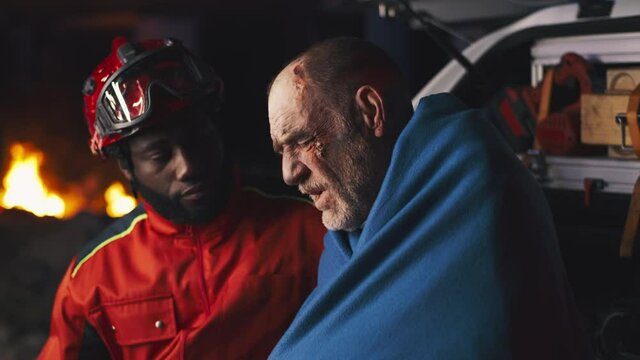 African American bearded man in paramedic uniform asking questions to senior man wrapped in blanket after accident at night