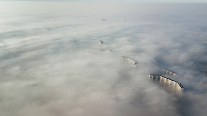Silhouettes of tall houses through the incoming thick fog. Megalopolis in white clouds in the early morning at time.