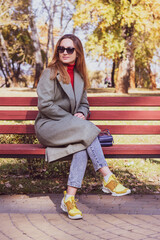 Red-haired girl in a coat sits on a red bench in autumn in the park