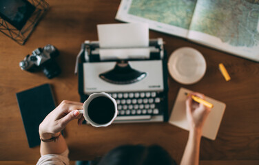 Fototapeta na wymiar High angle view of female writer's hand holding cup of coffee above desk