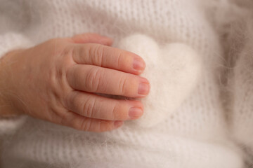 Tiny hand of a newborn baby. Soft hands of new born in white clothes hold white woolen heart. Fingers of a newborn baby hands close up. Studio macro photo. Happiness of a woman. Photography, concept.