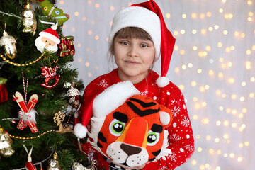 Close-up portrait of a cute Christmas girl in a red Santa hat and in a red Christmas sweater. Child stands at the Christmas tree with hand made toy tiger. Background of defocused lights.