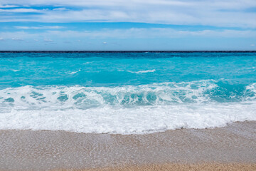 Fototapeta na wymiar Azure vibrant waves with white foam and blue sky on sunny coast of Greek island. Sandy beach in Greece. Summer vacation to Ionian Sea