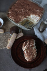 A piece of Tiramisu cake in a light plate on the table with a spruce towel and spoons. Dessert with mascarpone and savoyardi cookies.