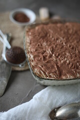 Tiramisu cake in a crystal square large plate on the table with a spruce towel and spoons. Dessert with mascarpone and savoyardi cookies.