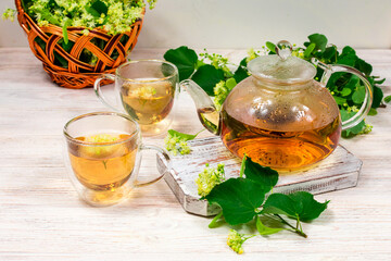 A teapot and two cups of tea with a linden tree on a wooden table against the background of a basket with linden flowers. Healing tea.