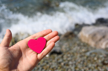Fototapeta premium Red heart on a man's palm against the backdrop of a pebble beach and sea water.
