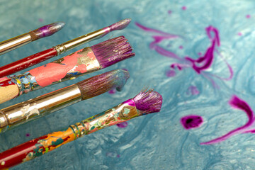 Photograph of five brushes of different thicknesses stained with metallic and shiny blue paint on a bucket of paint