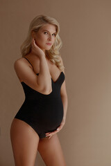 Fototapeta premium Adorable pregnant woman with blond hair in black bodysuits posing on the beige background