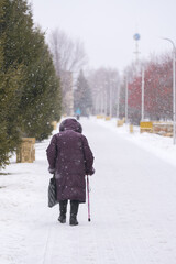 An elderly woman, leaning on a cane, carefully walks along the alley. Ice. Blizzard. Selective focus.