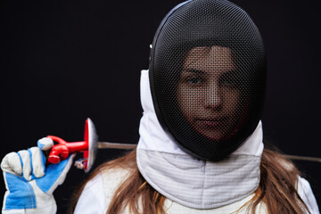 Portrait of a young fencer wearing white fencing costume and mask and holding the sword in front of...