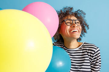 Fototapeta na wymiar Close up of happy multiracial lady looking away and smiling while holding birthday party balloons. Isolated on blue background