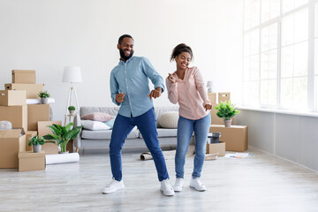 Smiling millennial african american husband and wife dancing together in room with cardboard boxes...