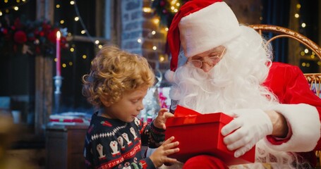 Santa Claus and little caucasian boy standing nearby and waiting for magical Christmas gift near the Christmas tree in cozy living room in winter