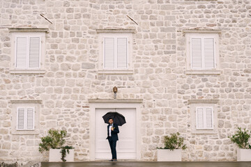 Groom stands under an umbrella near an old stone building with white shutters