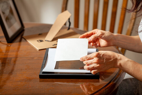 A young caucasian woman is placing a printed photograph into a picture frame with kickstand. There are other frames on the table. Tabletop picture framing concept.