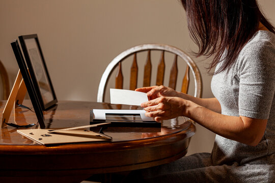 A young caucasian woman is placing a printed photograph into a picture frame with kickstand. There are other frames on the table. Tabletop picture framing concept.