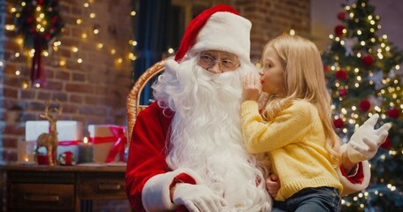I will tell you but keep it in secret. Portrait view of the little blonde girl sitting at the Santa knees while sharing secrets with him