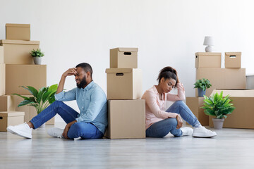 Tired upset young black husband and wife among cardboard boxes with things and plants in empty room