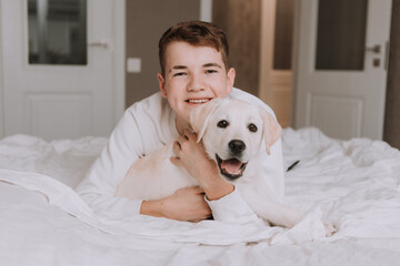 portrait of a teenage boy lying in bed on white bedding in an embrace with a light-colored dog. boy is fooling around with a pet in the room. space for text. High quality photo