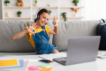 Girl sitting on couch, having video call wearing headphones waving