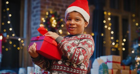 Little multiracial boy wearing Santa hat holding gift box with present while spending time at home with New Year decorations. Christmas spirit concept