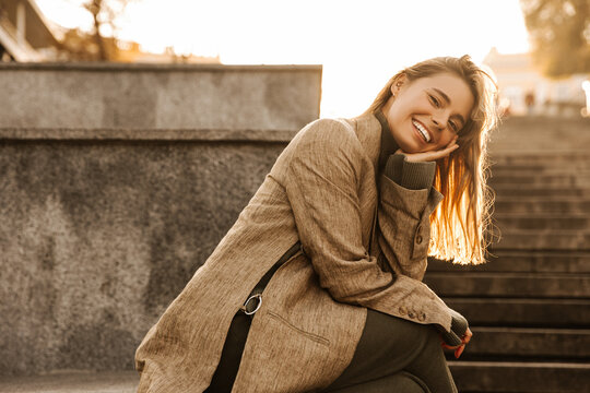 Image of beautiful stylish young caucasian woman sitting on street staircase with legs crossed on sunny day. Model resting head on her hand smiling looks at camera. Lifestyle concept