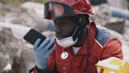 Black man in red uniform recording audio message on cellphone while sitting on rubble of demolished...
