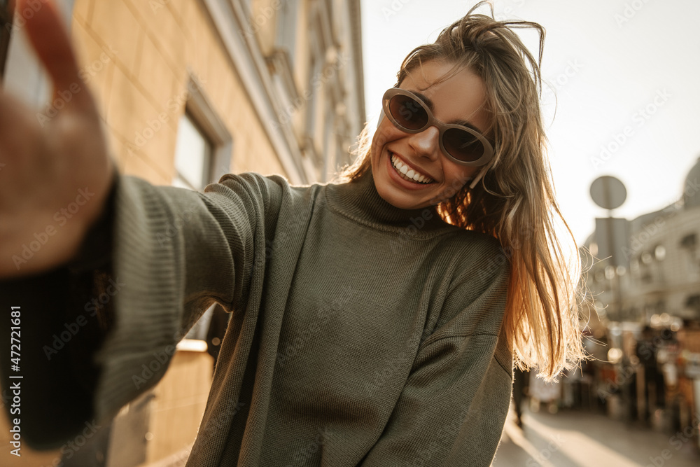 Wall mural Cute fair-skinned young woman makes happy selfie while holding camera with hand on background of city. Blonde in sunglasses is smiling broadly, wearing grey sweater. Lifestyle, female beauty concept