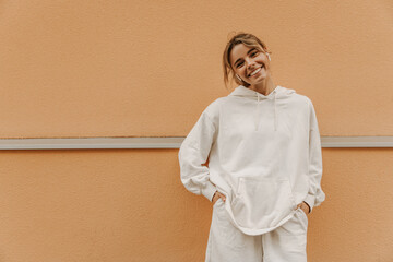 Beautiful caucasian young girl smiling with teeth looking at camera on background with place for text. Swarthy blonde beauty wears white warm tracksuit. Good mood, fashion trends