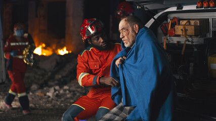 African American bearded man in paramedic uniform asking questions to senior man wrapped in blanket...