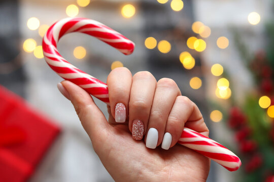Woman's hand with gel polish manicure white color and with snowflakes ornament, holding candy cane against festive Christmas background. Selective focus. Idea of the winter manicure