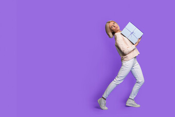 Side view portrait of a caucasian woman standing and trying to hold heavy gift box looking at camera. Facial expressions. Purple background with copyspace.