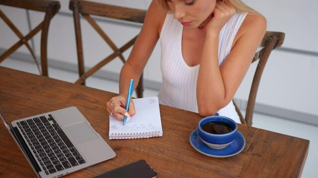 Young student girl in a white top working from home sits at the table and makes notes during vebinar