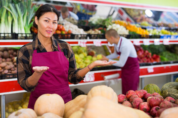 Smiling Asian saleswoman inviting to fruit and vegetable store, offering fresh organic produce..