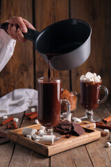 Woman pouring hot chocolate from pot into glass cup on wooden background