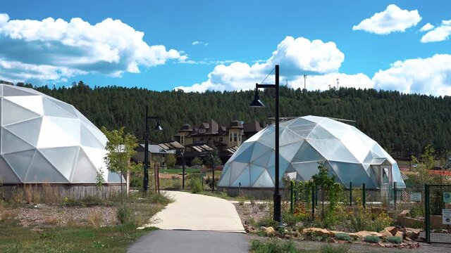 Wide shot of futuristic Geodisic Dome Greenhouses in rural area during sunny day - overgrown mountains in background