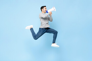 Portrait of energetic young Caucasian man jumping with megaphone isolated on light blue background