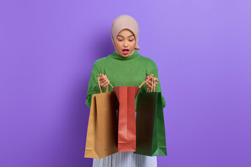 Shocked beautiful Asian woman in green sweater opening shopping bags isolated over purple background