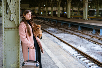 Beautiful young casual tourist woman with dog and suitcase waiting for train at train station....