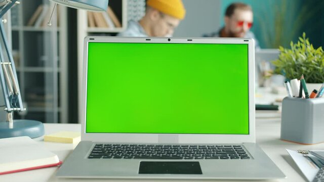 Chroma key green screen laptop on desk with employees moving in background in office. Modern technology and online content concept.