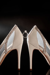 Beautiful white bride's shoes and golden wedding ring on dark table background, copy space. Wedding concept
