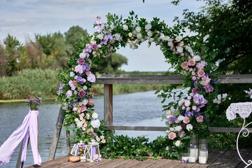Circle wedding arch decorated with white, pink and purple flowers and greenery on terrace by lake...