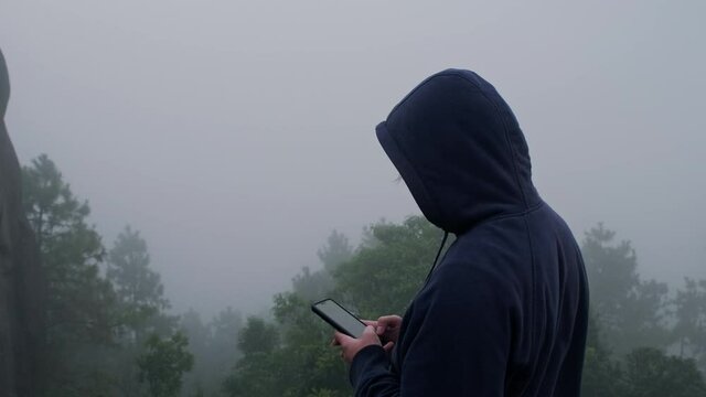 Young man traveler in navy blue male hoodie sweatshirt long sleeve using a smartphone and enjoy the scenery in the mountains with thick fog in the background.