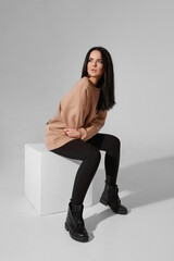 Adorale young woman with trendy makeup wearing trendy sweatshirt sitting on the white cube over the white background